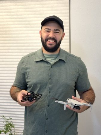 A smiling man with a beard and black cap holds two drones—an FPV-style cinewhoop in his right hand and a DJI Mini 2 in his left hand. He is wearing a green button-up shirt and standing indoors in front of a light-colored window shade.